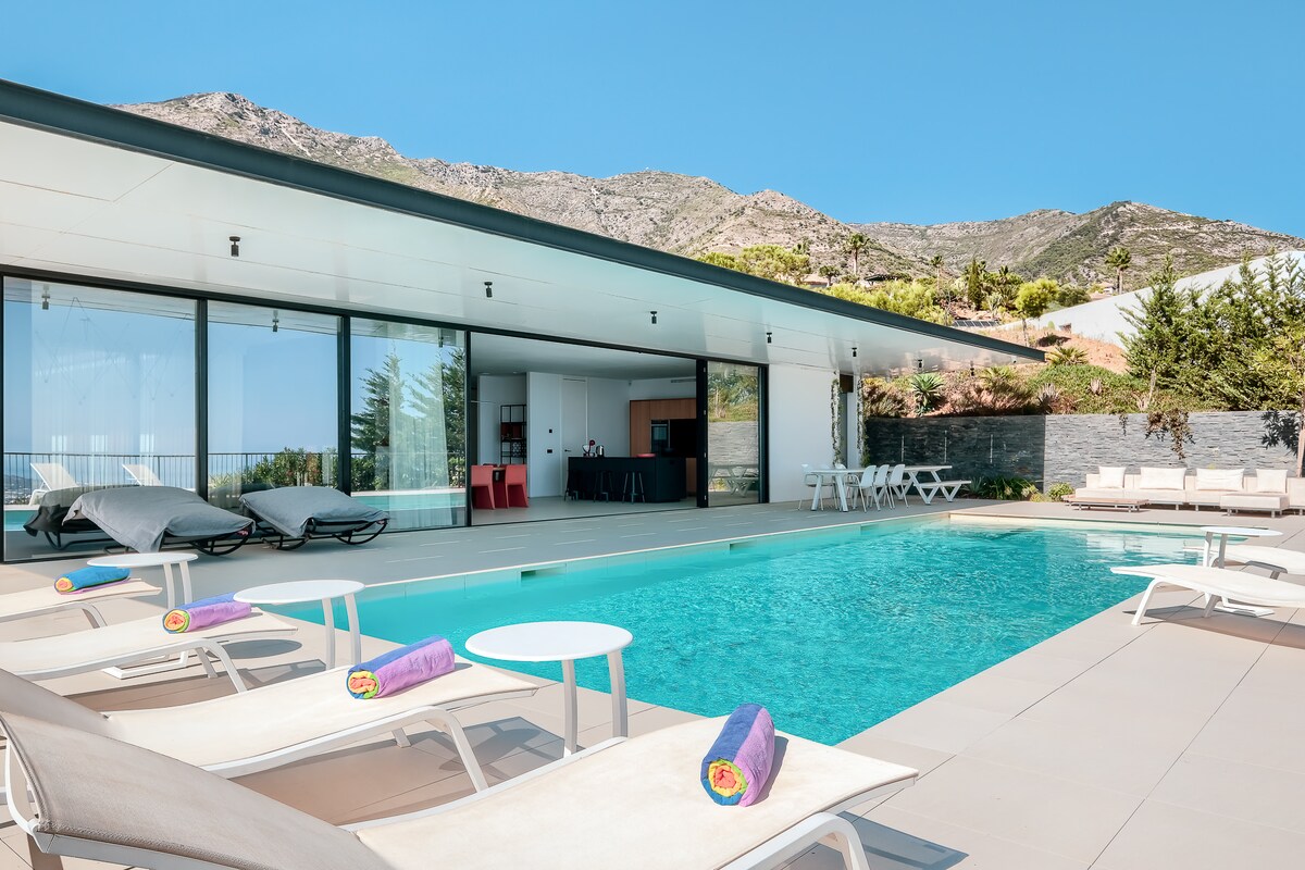 An inviting outdoor area features a turquoise swimming pool surrounded by modern lounge chairs, each with colorful towels. The sleek villa exterior is visible in the background, showcasing large glass doors that offer unobstructed views of the surrounding mountains.