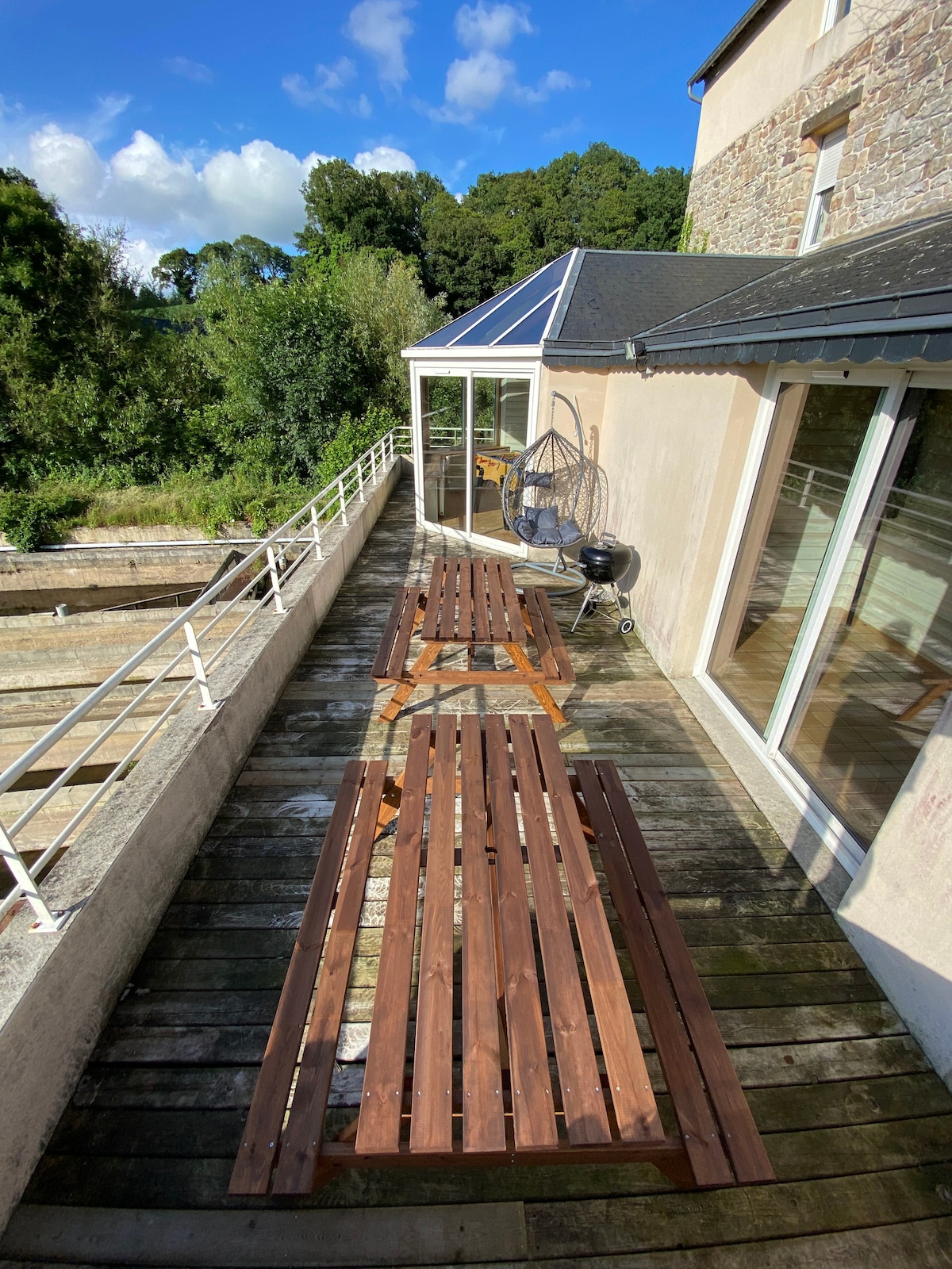 A spacious outdoor terrace is highlighted, featuring two wooden tables arranged for dining. A cozy seating area is visible, accompanied by lush greenery that frames the surrounding landscape. Large glass doors lead into the interior, allowing natural light to flood the space.