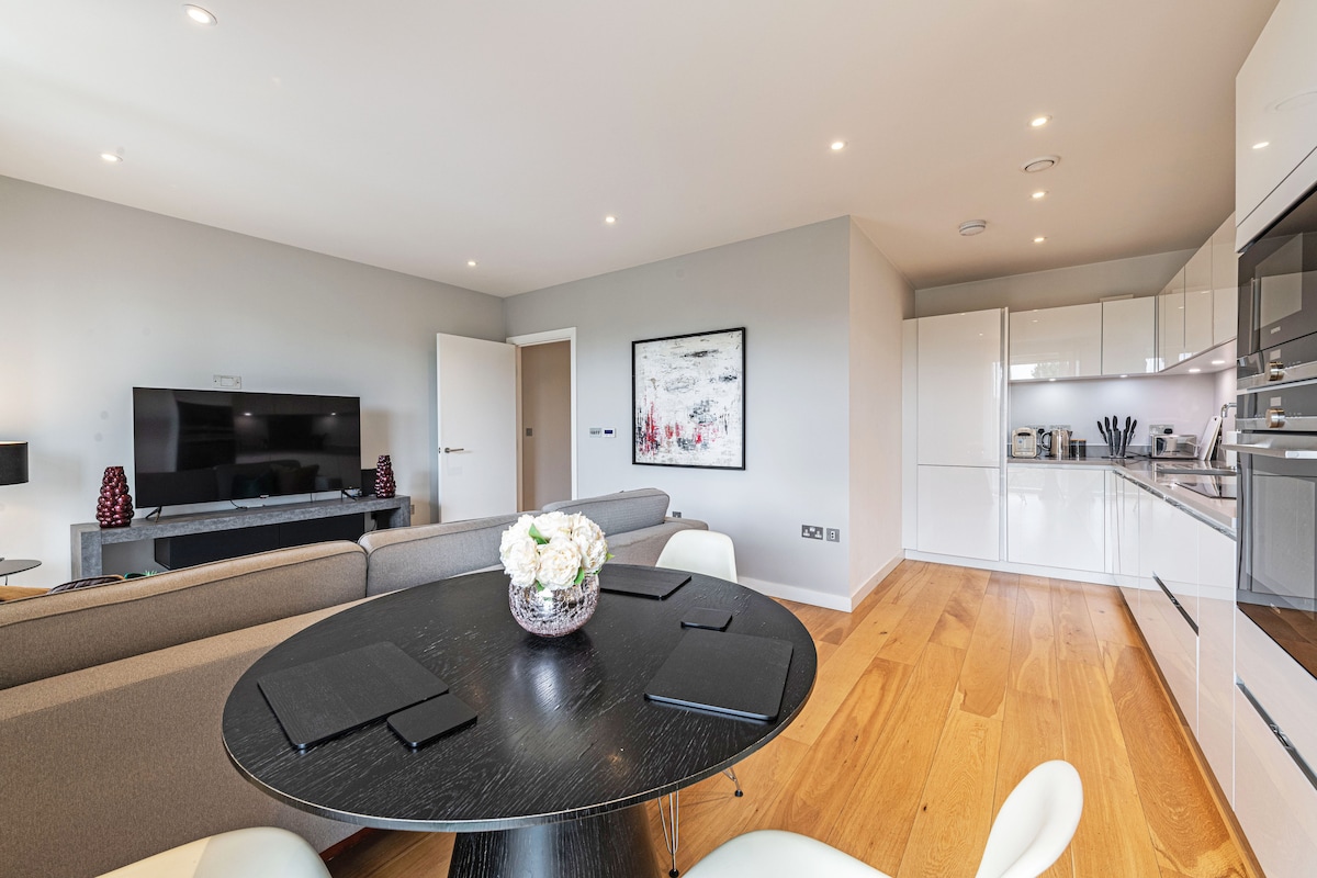 A modern living area showcases a sleek gray sofa and a round black dining table set for four. The open kitchen features glossy white cabinetry and stainless steel appliances. A contemporary piece of wall art adds a subtle touch of color to the neutral space.