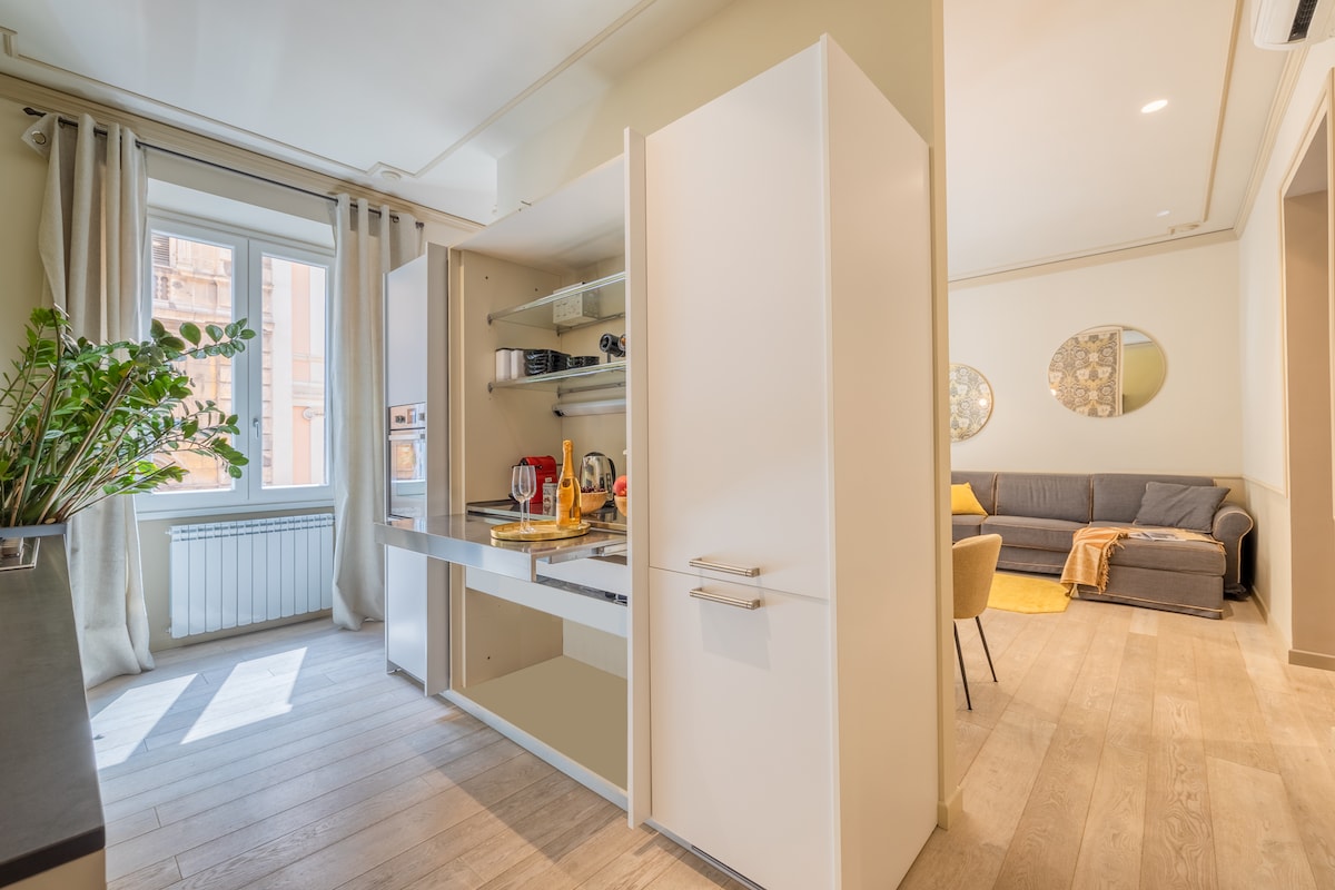 A modern kitchen area is visible, featuring sleek cabinetry and an island equipped with various kitchen appliances. Natural light is filtered through a large window, highlighting the open layout that connects to a comfortable living space furnished with a gray sofa.