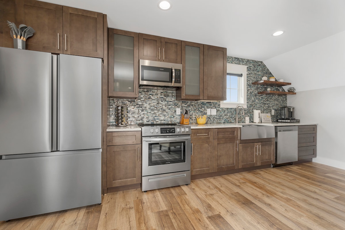 A modern kitchen features stainless steel appliances, including a refrigerator, oven, and microwave. Dark wood cabinetry provides storage alongside a textured backsplash. A sink and dishwasher are integrated within the counter. Natural light fills the space through a window, highlighting the warm wood flooring.