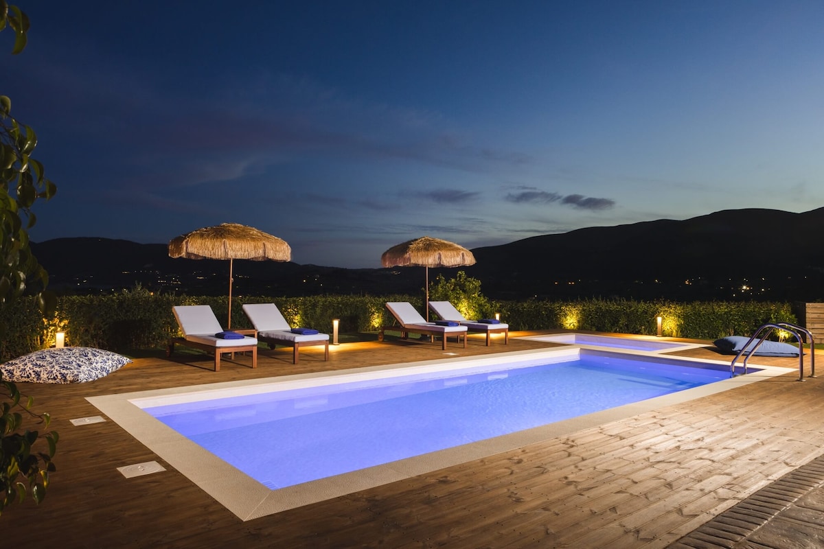 An outdoor pool is softly illuminated, bordered by wooden decking. Sunbeds are neatly arranged under thatched umbrellas, offering a serene space for relaxation. Lush greenery surrounds the area, creating a tranquil view of the distant hills against the night sky.