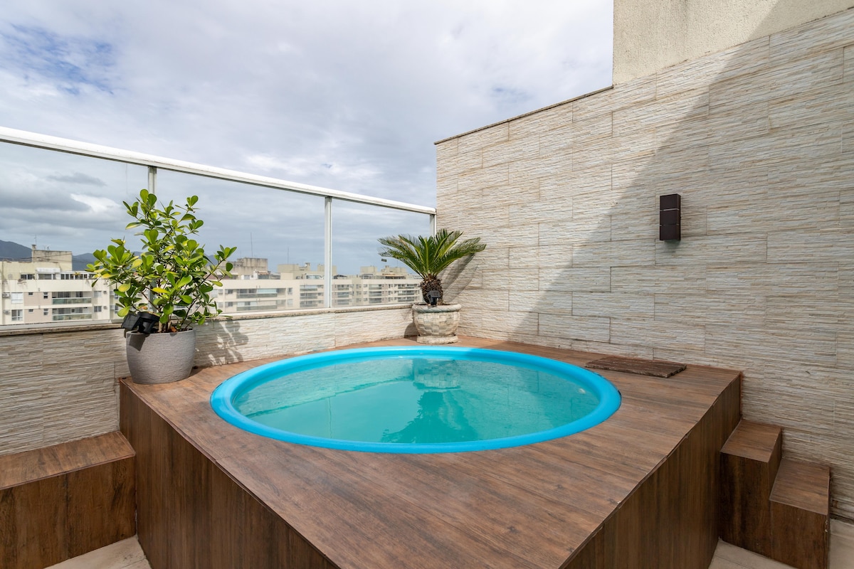 A circular hot tub is presented against a backdrop of expansive city views. The wooden deck surrounds the tub, while decorative plants add a touch of greenery. Glass railings provide a sense of openness and safety.