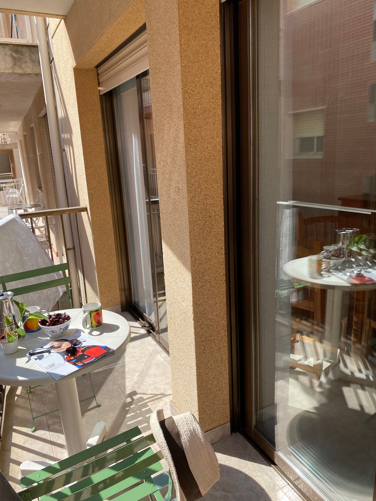 A small balcony area features a round table with two green chairs. A glass of refreshing drink and a plate of fruit are placed on the table. Natural light illuminates the space, highlighting the warm tones of the textured walls and the nearby windows.