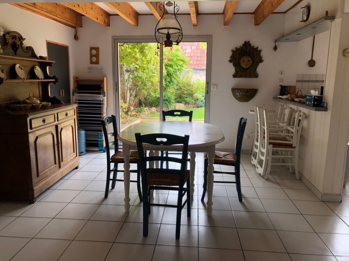 A dining area features a round white table surrounded by chairs in various colors. A wooden sideboard is positioned against the wall, and a sliding glass door offers a view of a green outdoor space. Light-colored tiles cover the floor.