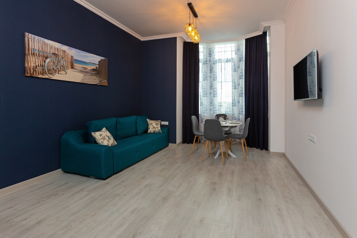 A living area is depicted with a deep blue accent wall. A plush, teal sofa is positioned against the wall, complemented by decorative pillows. A dining table with four gray chairs is situated nearby, illuminated by natural light streaming through large windows adorned with sheer curtains.