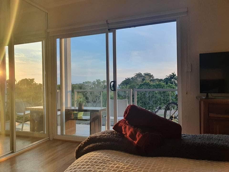 The studio features glass sliding doors that open to a balcony, providing views of the sunset and surrounding greenery. A spacious bed is adorned with a maroon throw, positioned near a wooden cabinet and a television. Soft evening light enhances the warm atmosphere.