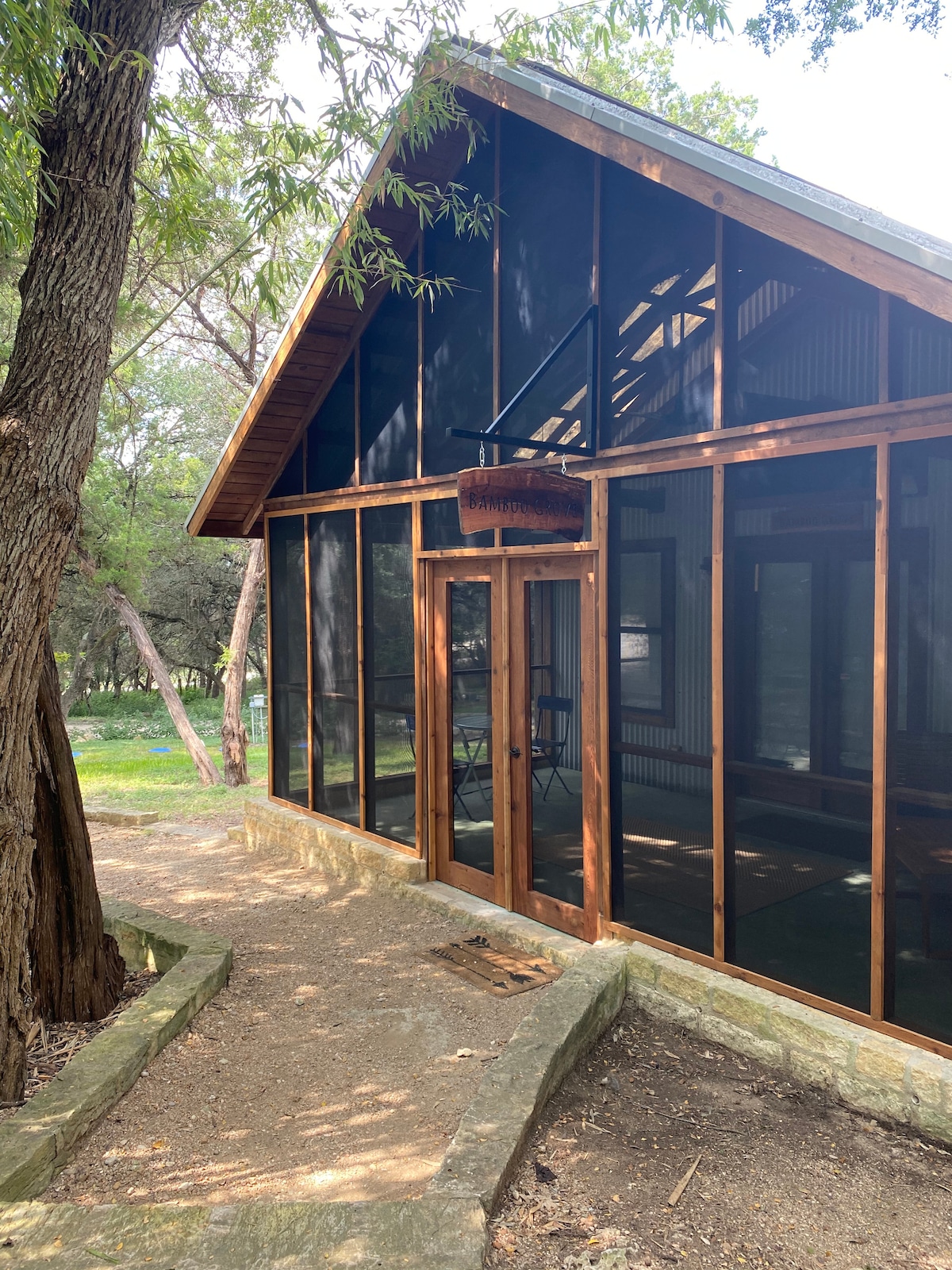A cabin entrance is framed by large glass panels and wooden beams, allowing natural light to filter in. The door is surrounded by nature, with a pathway leading to the porch. Shade from nearby trees adds a serene atmosphere.