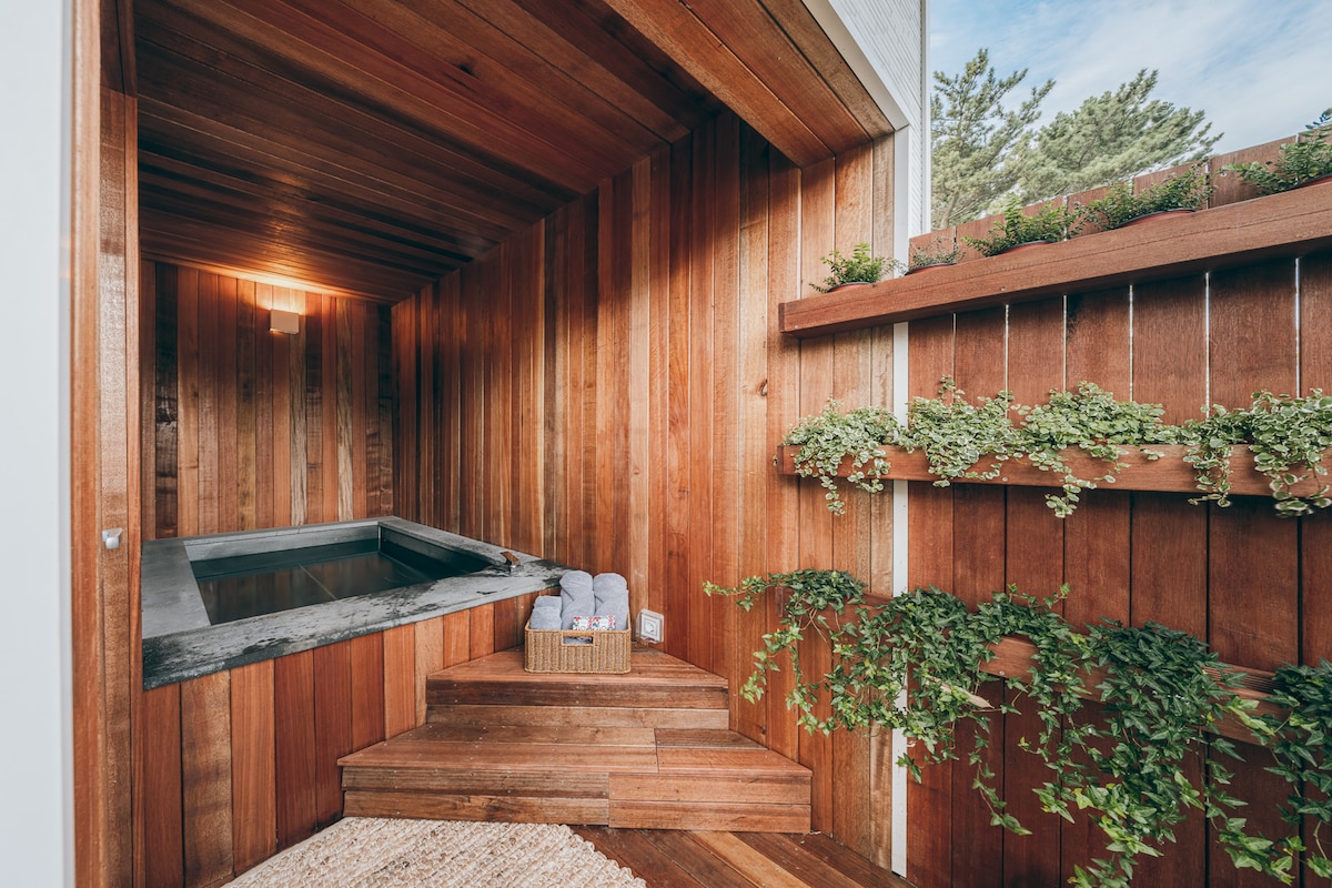 A wooden sauna space features natural wood paneling and a stone-lined jacuzzi. A simple basket holds rolled towels on a step, while greenery adorns the walls. Soft lighting illuminates the area, creating a calming environment for relaxation.