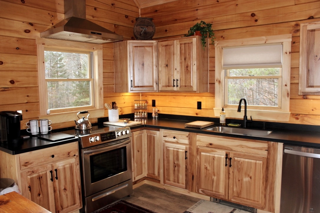 A functional kitchen design features natural wood cabinetry paired with a dark countertop. Modern stainless steel appliances are prominently displayed, including an oven and dishwasher. Two windows allow for natural light, while a kitchen sink and various utensils are arranged thoughtfully for convenience.