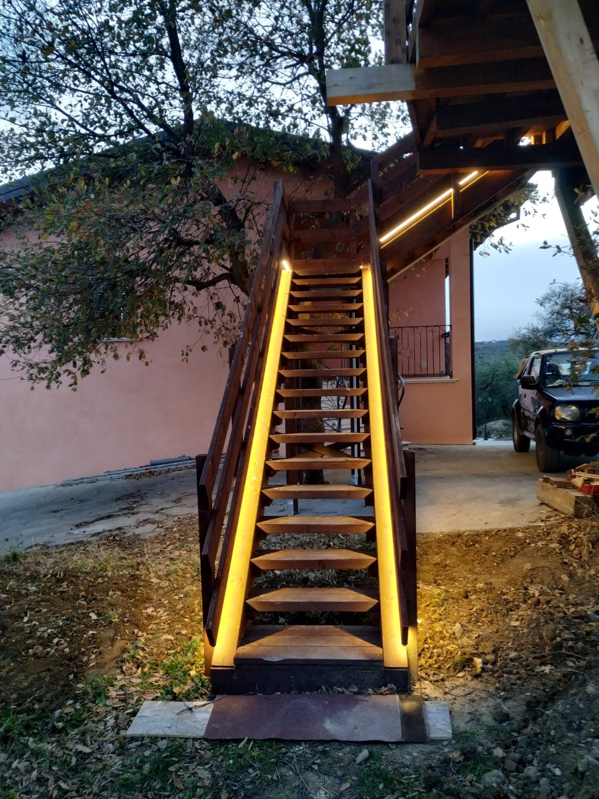A wooden staircase, gently illuminated by soft lighting along the edges, leads to the elevated entrance of the treehouse. Surrounding greenery and a glimpse of a parked vehicle are visible, providing a natural setting for the structure.