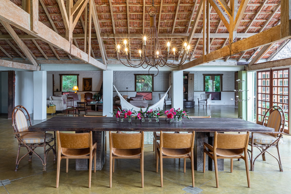 A spacious dining area is highlighted by a large wooden table surrounded by several tasteful chairs. Exposed wooden beams and a decorative chandelier add character to the open space, which seamlessly connects to comfortable seating areas and large windows providing natural light.