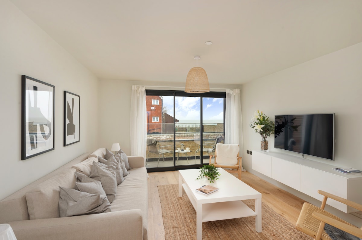 A light-filled living area features a comfortable sofa adorned with various textured cushions. Large glass doors provide a view of the outdoor terrace and sea, while a sleek white coffee table sits in front of a modern entertainment unit. Decorative elements enhance the inviting space.