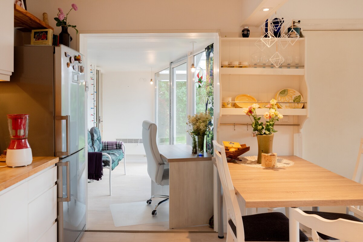 A modern kitchen space is featured, showcasing a refrigerator and a wooden countertop. A dining table is set with floral arrangements. Natural light enters through large glass doors, illuminating a nearby seating area with green accents and a desk for potential workspaces.