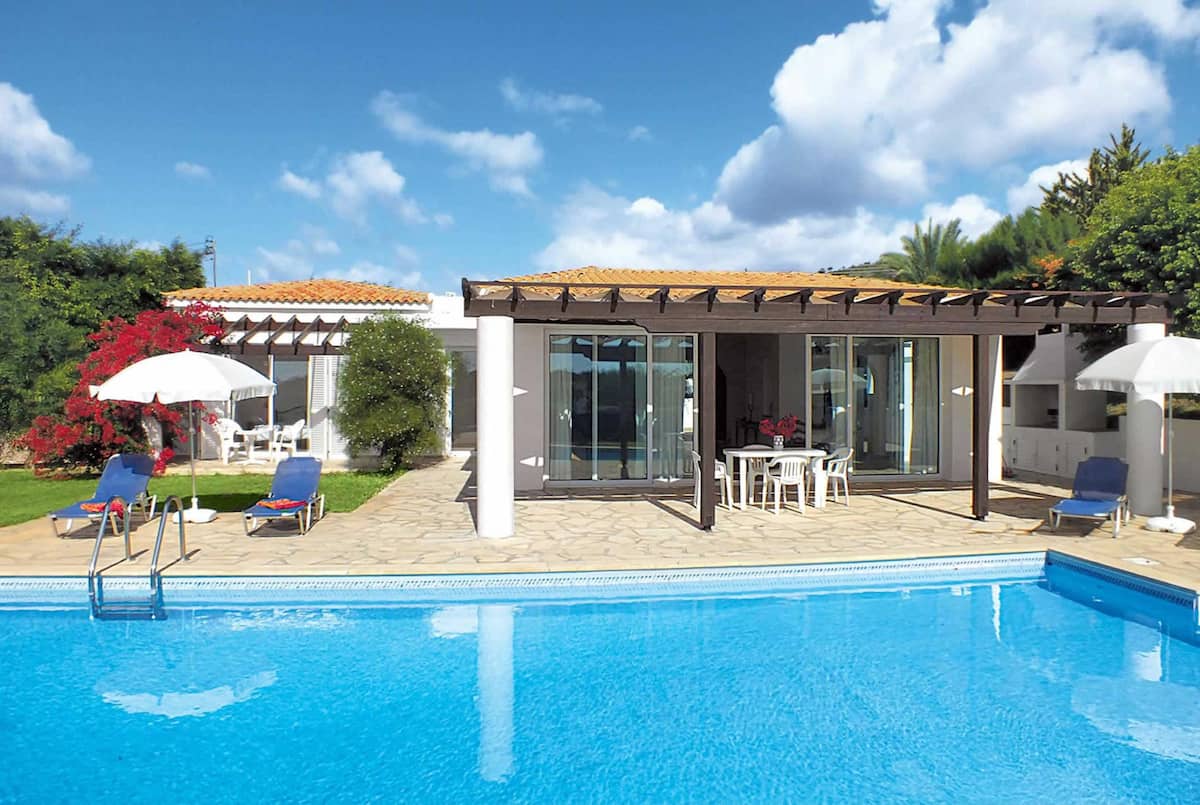 A villa exterior is shown, featuring a spacious swimming pool surrounded by lounge chairs and umbrellas. Sliding glass doors provide access to the indoor living area, while vibrant flowers and greenery enhance the outdoor space. The sky above is partly cloudy.