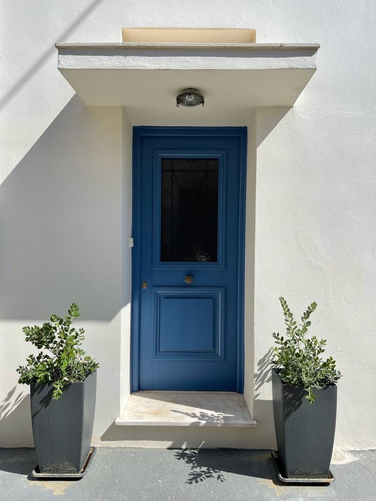 A blue door is framed by a light-colored wall, featuring a small overhang above. On either side, tall black planters with green foliage are positioned, adding a touch of nature to the entrance.