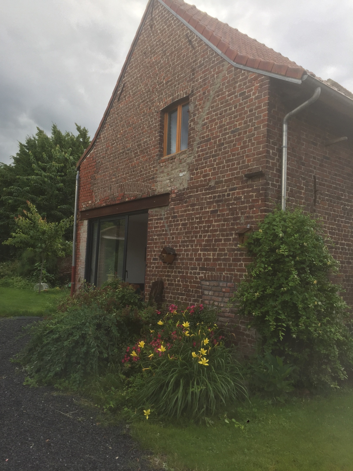 A rustic exterior of a brick building is visible, featuring a large window framed by leafy greenery. Colorful flowers bloom in the garden area, enhancing the natural surroundings. The structure showcases a pitched roof, blending harmoniously with the tranquil landscape.