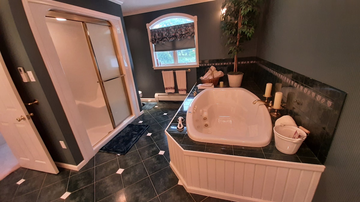 A spacious bathroom features a large garden tub alongside a separate glass-enclosed shower. The dark green tile flooring complements the light-colored walls. Soft lighting illuminates decorative elements, including candles and a small plant. Towels are neatly arranged on a wooden rack near the window.