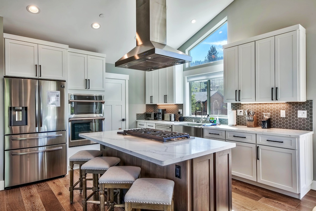 A modern kitchen is presented with stainless steel appliances, including a refrigerator and built-in oven. A large quartz island with seating accommodates gatherings. Windows above the sink provide natural light, and a stylish range hood hovers above the gas stovetop, enhancing the functional layout.