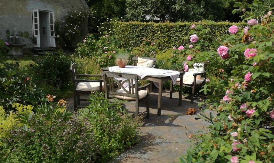 An outdoor seating area is surrounded by lush greenery and blooming roses. A wooden table is complemented by several chairs, providing a cozy space for gatherings. Stone paving creates a sturdy surface, while an inviting garden atmosphere is enhanced by the vibrant flowers.