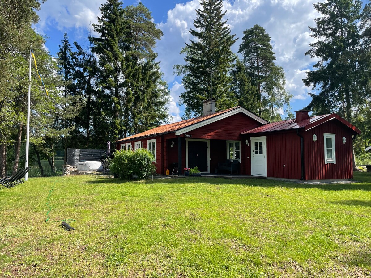 A charming red cabin stands amidst tall trees and green grass, featuring a welcoming porch and a small backyard area. A lazy spa can be seen on the side, complemented by a hammock for relaxation. Sunlight filters through the scattered clouds, illuminating the scene.