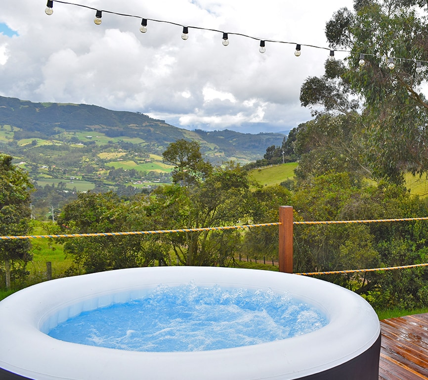An inflatable hot tub is set on a wooden deck, surrounded by lush greenery and expansive mountain views. Soft lighting from hanging strings illuminates the area, creating a serene environment for relaxation amidst nature.