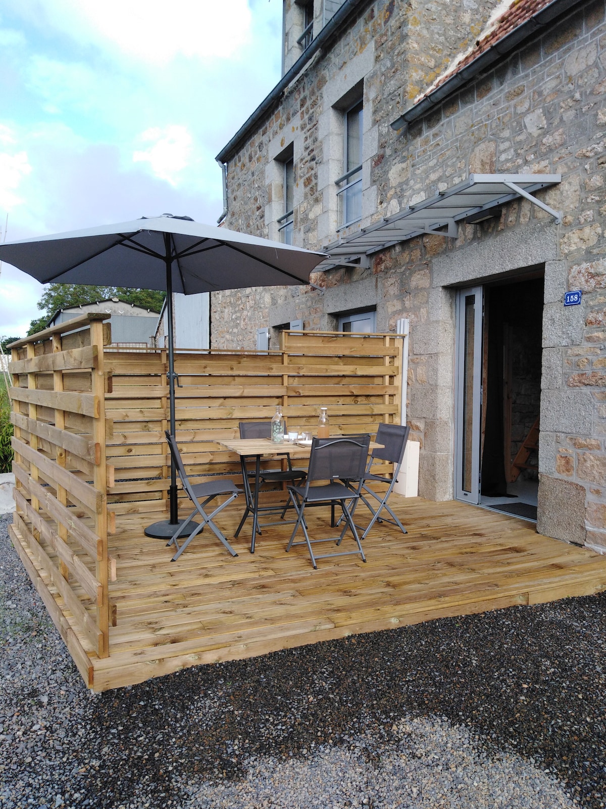 A wooden terrace is featured, equipped with a table and four chairs. A large umbrella provides shade over the dining area, while wooden slats form a partial enclosure around the space. The entrance to the house is visible nearby.