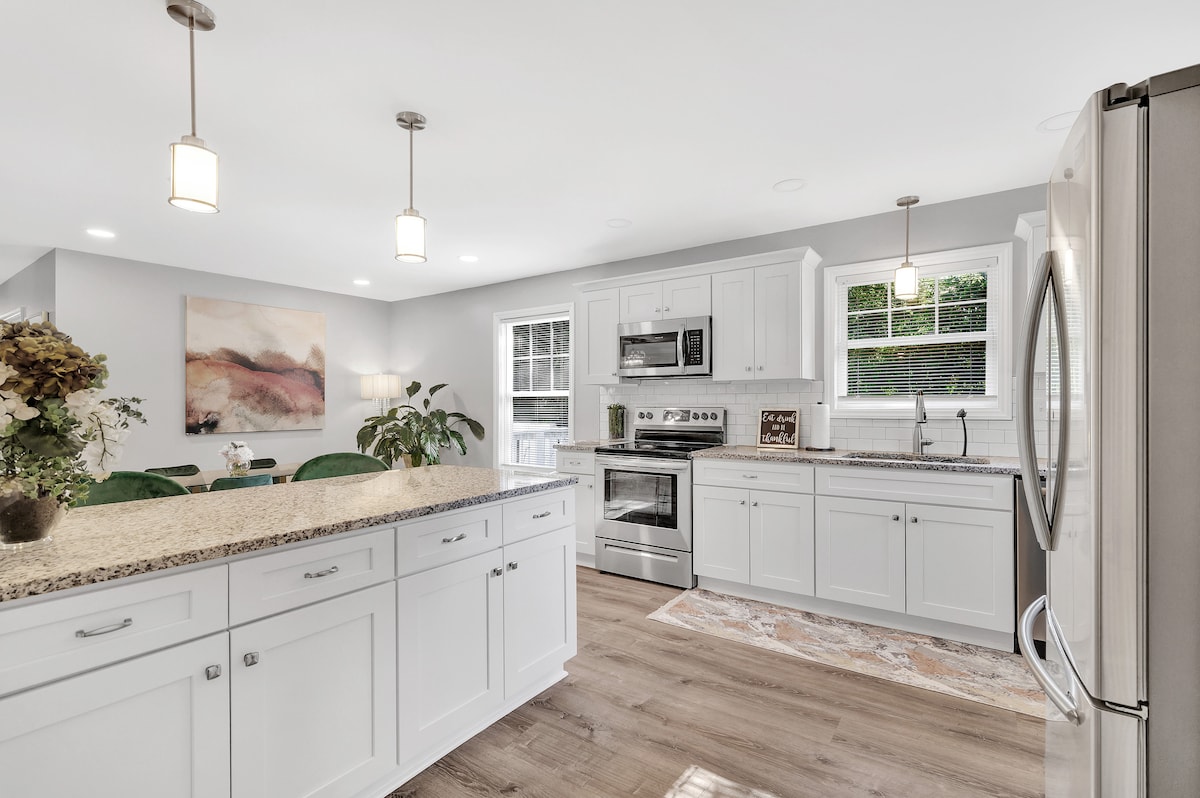 A modern kitchen features white cabinetry and granite countertops. Stainless steel appliances, including a microwave and oven, are visible. Natural light filters through the windows, illuminating the space. A dining area is accessible with green chairs and decorative plants enhancing the ambiance.