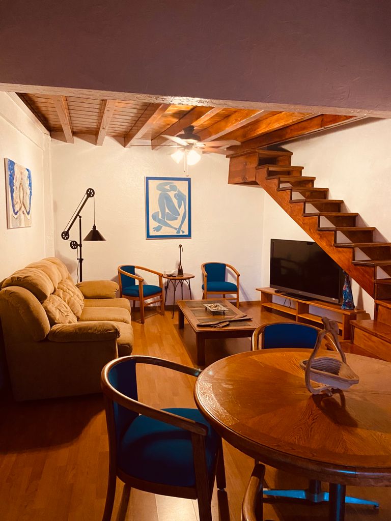 The living area features a comfortable sofa, two blue chairs, a wooden coffee table, and a television. A staircase leads to the upper level, while art pieces hang on the walls, contributing to the space's unique character. Natural light is provided by a ceiling fan.