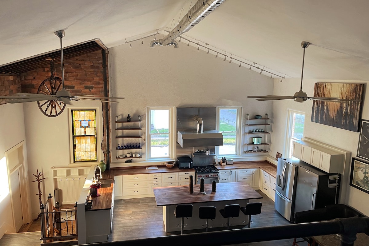 The spacious kitchen features high ceilings and large windows that allow natural light. A long wooden island sits at the center, flanked by three bar stools. Modern appliances, including a stainless steel refrigerator and stove, are positioned along the perimeter, complemented by wooden shelves and decorative elements.