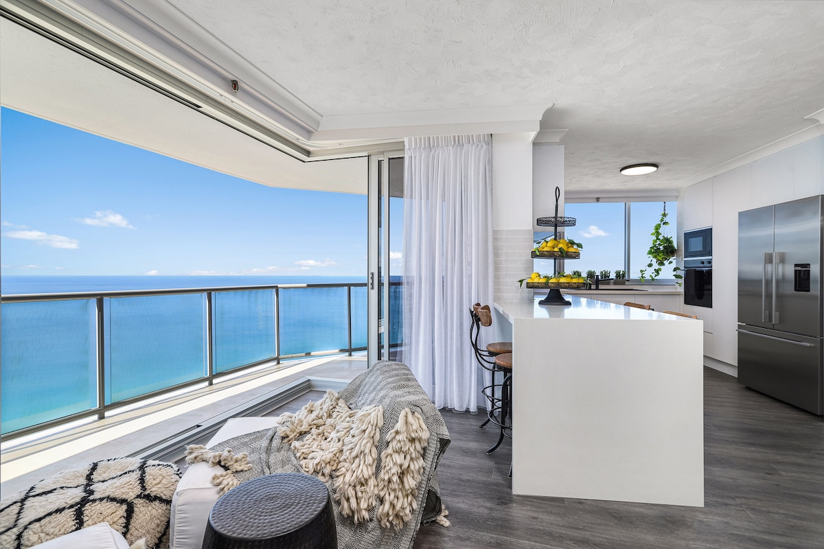 A bright and modern kitchen area features a large island with seating, surrounded by floor-to-ceiling windows offering expansive ocean views. Light drapes softly frame the ocean backdrop, while a plush, textured throw is draped casually over a lounge chair.
