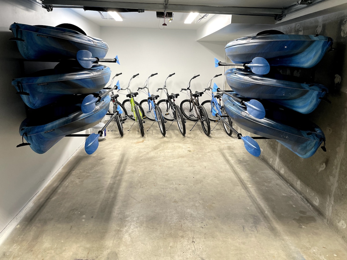 A spacious garage area is presented, featuring six kayaks securely mounted on one side and six cruiser bikes lined up on the opposite side. The concrete flooring offers a neutral backdrop, contributing to an organized and functional storage space.