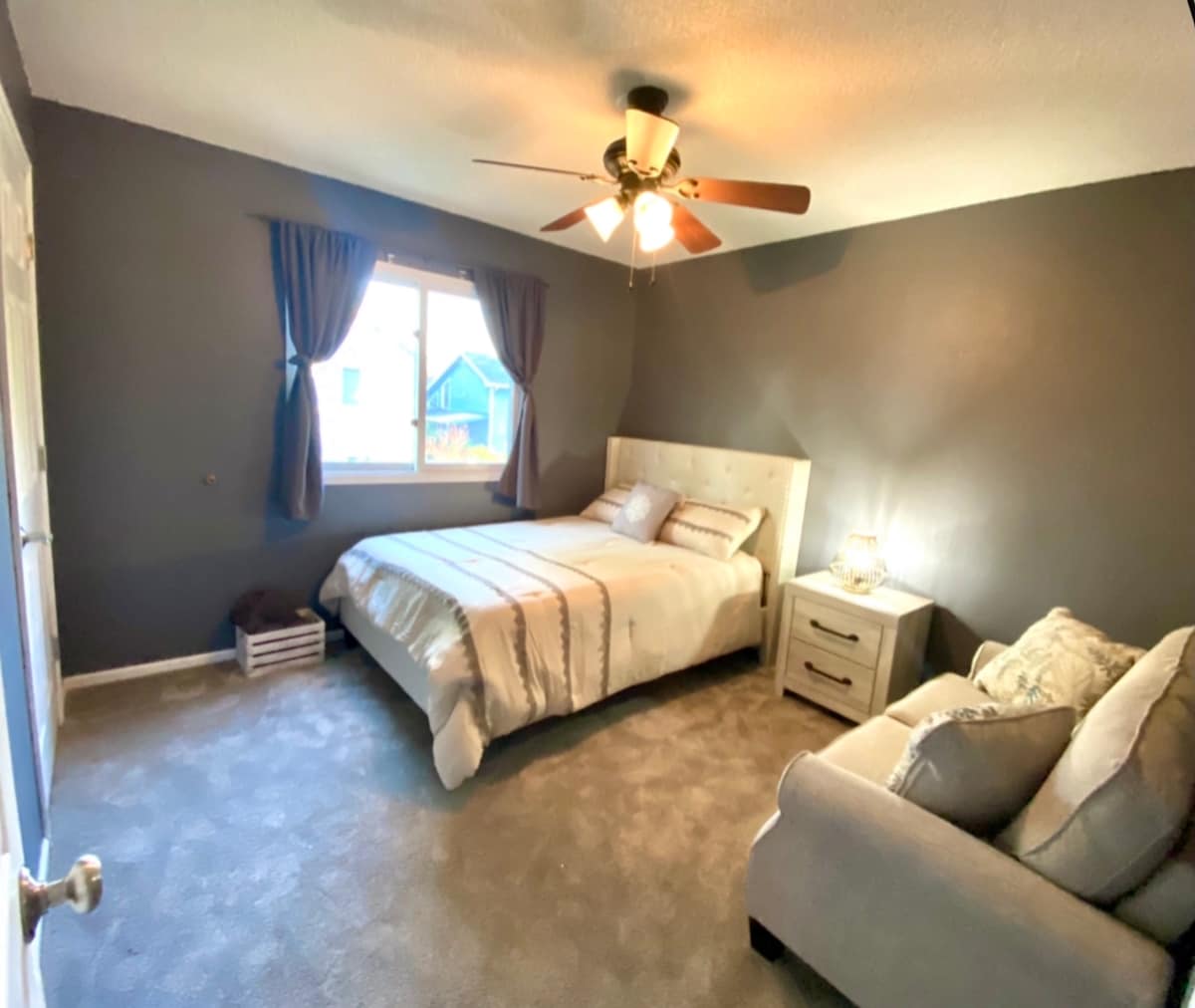 A serene bedroom is presented with dark gray walls and a spacious bed with light bedding. A window allows natural light to enter, and a cozy couch is positioned beside the bed. A nightstand holds a lamp, adding functionality to the space.