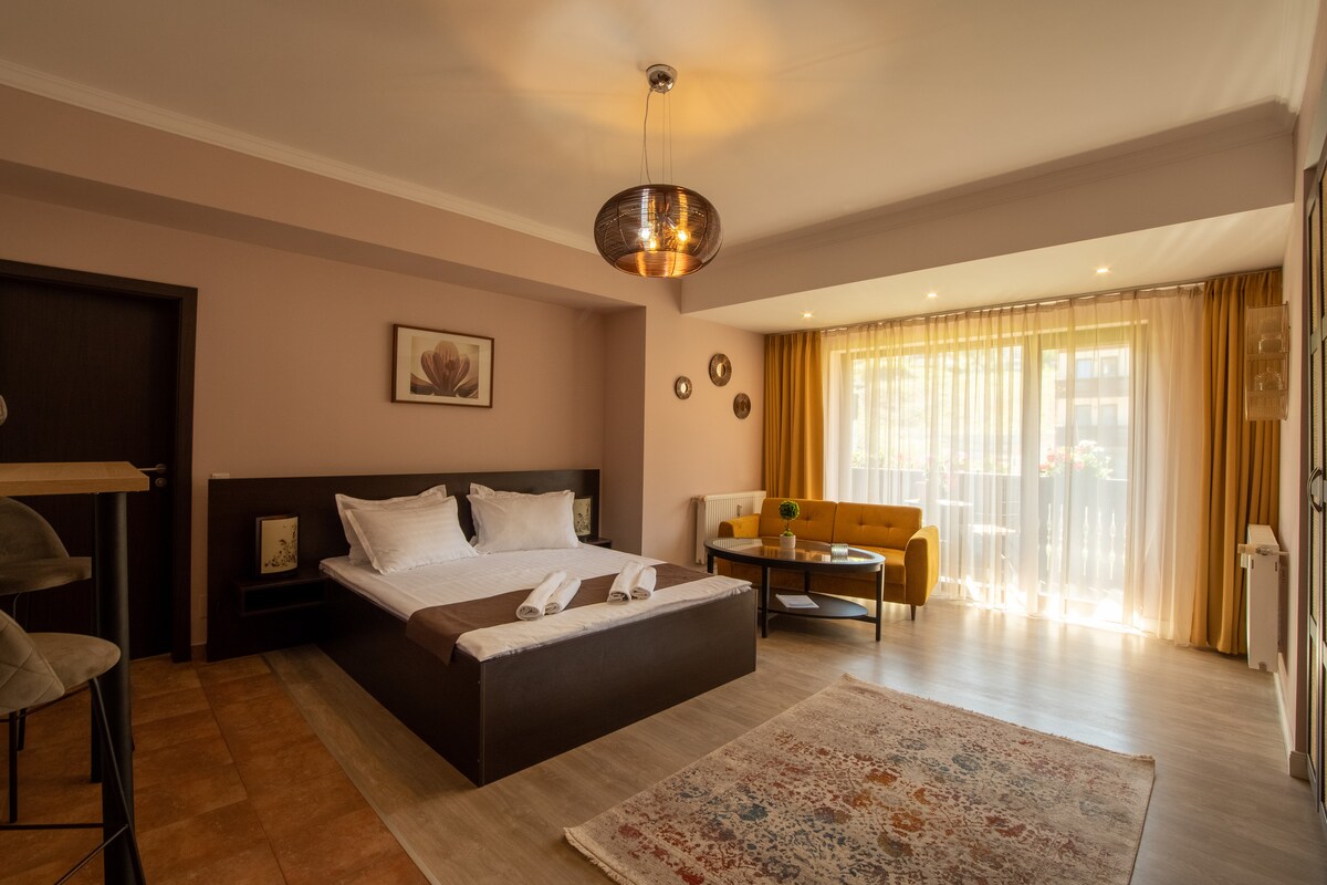 A spacious studio is highlighted by a king bed with neatly arranged towels. Natural light floods through large windows, illuminating a cozy seating area with a yellow sofa and a circular coffee table. Decorative elements include a flower artwork and stylish lighting fixtures.