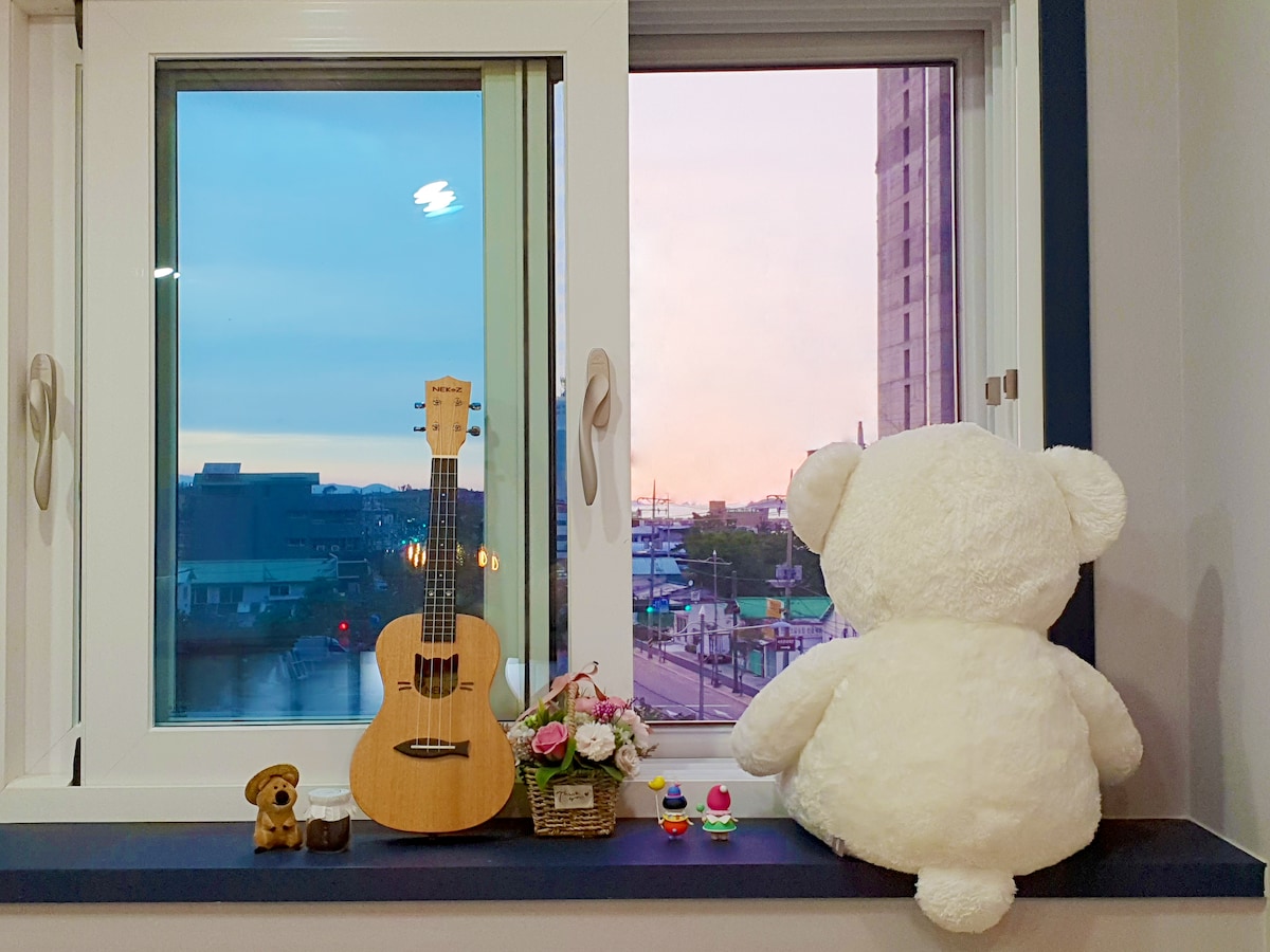 A window view shows a soft teddy bear gazing outside, accompanied by a small acoustic guitar and a decorative basket with flowers. The scene captures a pastel sky at sunset, enhancing the cozy atmosphere of the room.