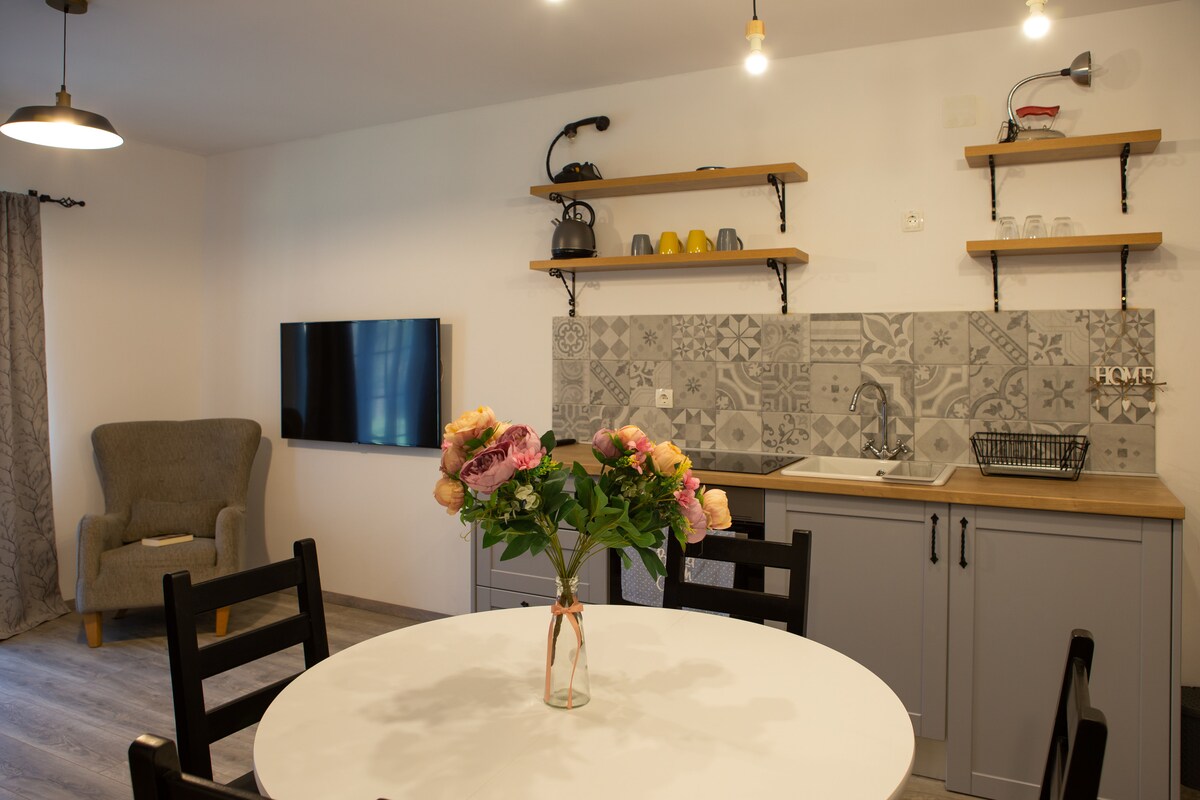 A modern kitchen area features gray cabinetry and open shelving displaying colorful dishware. A round dining table is set with a floral centerpiece. A cozy armchair is positioned nearby, and a flat-screen TV is mounted on the wall, enhancing the functional living space.