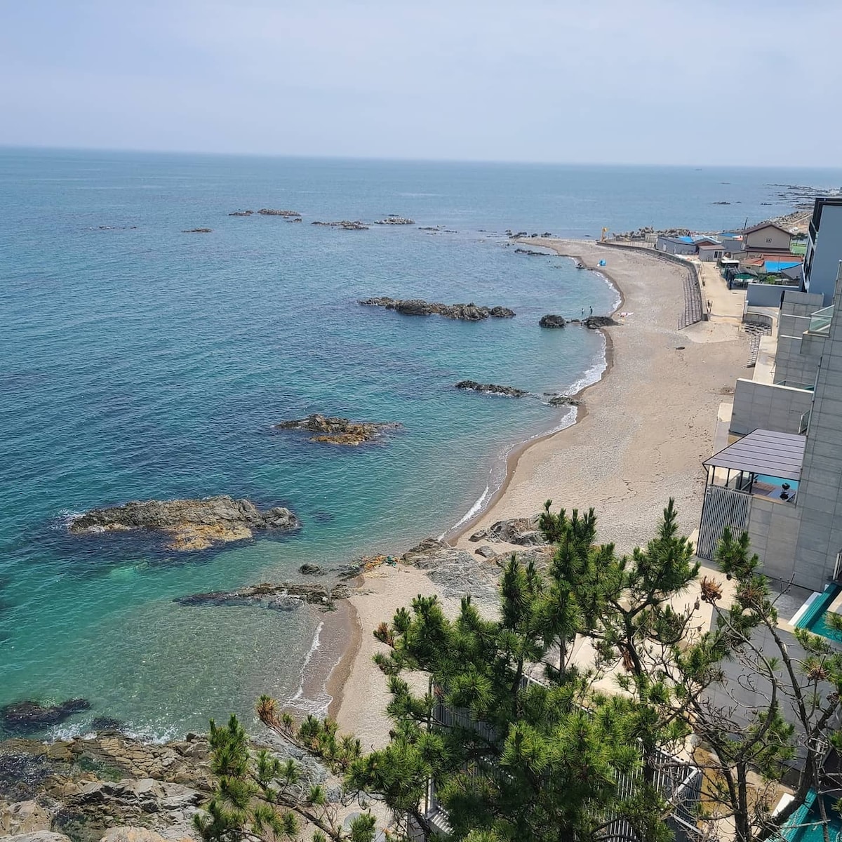 A serene coastal view features a sandy beach bordered by rocky formations. Gentle waves lap at the shore under a clear sky. In the distance, a few scattered structures hint at a tranquil beachfront atmosphere, providing an inviting space for relaxation and exploration.
