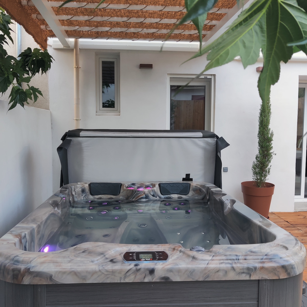 A private jacuzzi is positioned on a wooden deck, featuring a marbled surface and illuminated with soft purple lights. Green foliage from potted plants adds a natural accent, while a partially visible wall with windows creates a serene outdoor atmosphere.