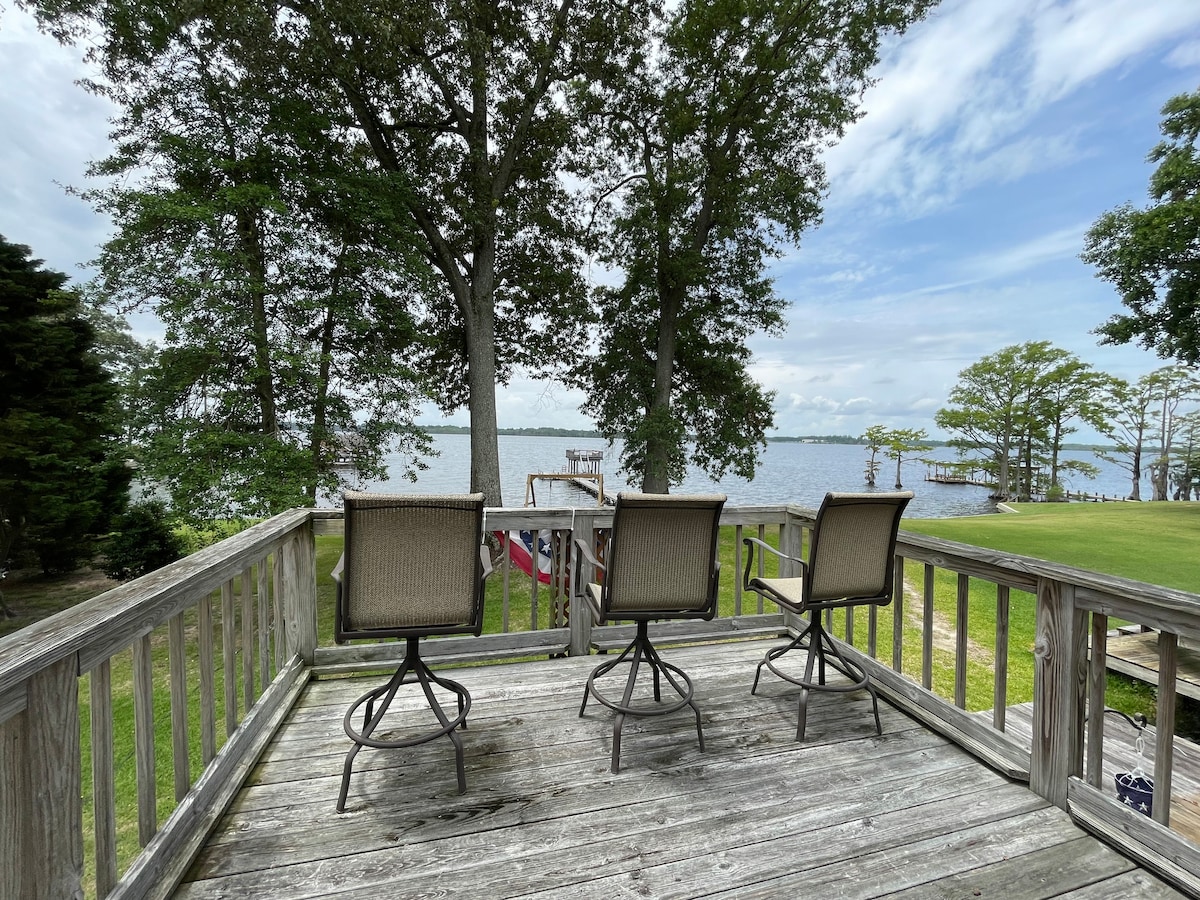 A spacious deck features three elevated chairs, offering views of the Perquimans River. Surrounding the deck are green trees, and the water sparkles in the sunlight. The scene creates an inviting spot for relaxation and enjoyment of the natural surroundings.