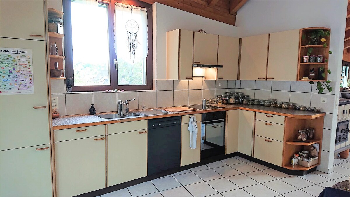 A well-equipped kitchen features beige cabinetry and a tiled countertop. Natural light enters through large windows, illuminating the space. Kitchen appliances, including a dishwasher and sink, are present, along with neatly arranged jars on shelves, enhancing the functionality of the area.