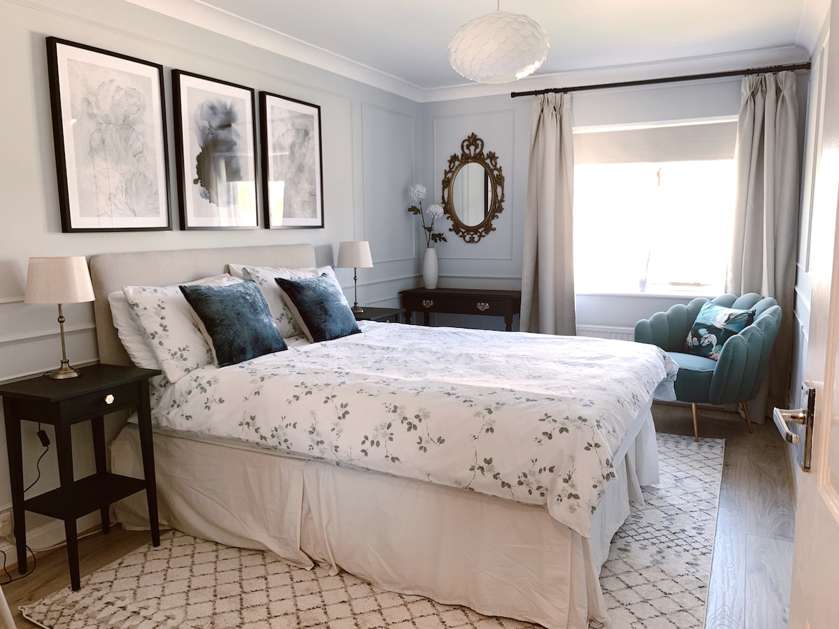 A bright bedroom features a spacious bed dressed in floral bedding. Two bedside lamps are positioned on either side. A decorative mirror reflects light from a window adorned with light curtains. A cozy chair in the corner complements the neutral color palette.
