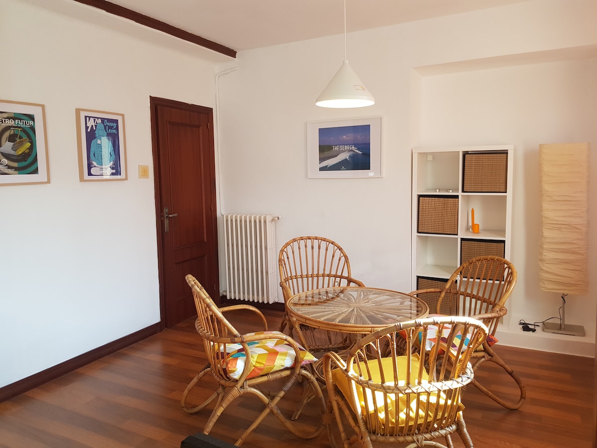 A spacious living area features a round glass table surrounded by four rattan chairs with colorful cushions. The room is illuminated by a pendant light, and framed artwork adorns the walls. A white shelving unit stands with woven storage baskets, contributing to the inviting layout.