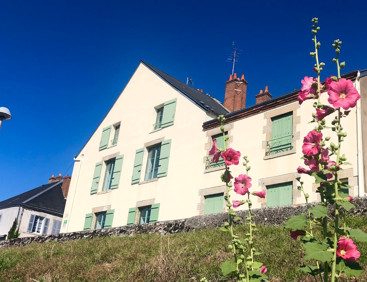 A charming house with light-colored walls and green shutters is set against a clear blue sky. The floral plants in the foreground add a touch of color, while multiple windows suggest natural light within the spacious interior.