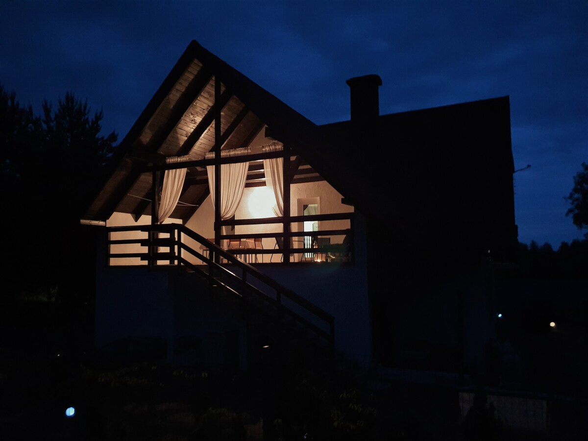 The exterior of the building is softly illuminated at night, highlighting the rustic architecture. A balcony with railing is visible, adorned with draped fabric. Surrounding greenery adds to the serene atmosphere, while the house maintains a welcoming presence against the evening sky.