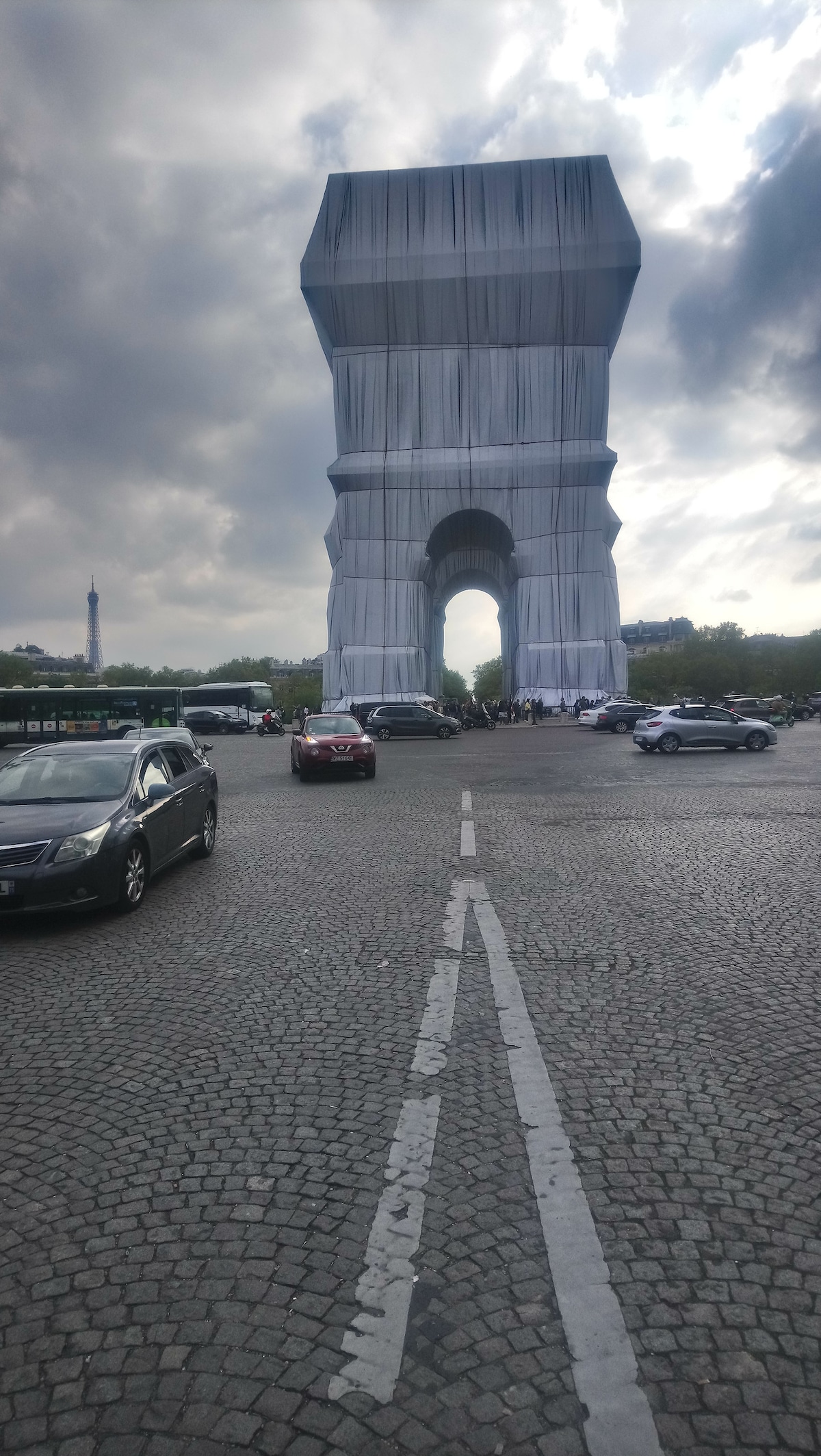 An iconic archway is displayed, covered in fabric, with vehicles surrounding it on a cobblestone road. The arch stands tall and is flanked by trees, with a cloudy sky above. The Eiffel Tower is visible in the distance, adding to the city's charm.