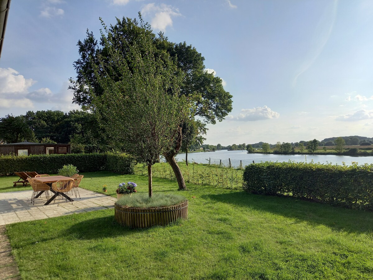 A scenic view of the garden is presented, featuring a neatly manicured lawn surrounded by hedges. A patio area with lounge chairs offers a relaxing space, while a tree provides shade. The nearby river reflects the sky, enhancing the tranquil outdoor setting.
