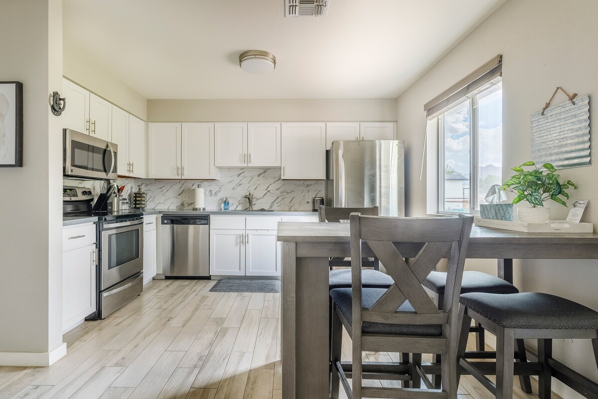 A modern kitchen features white cabinetry, stainless steel appliances, and a marble backsplash. A wooden dining table with grey chairs is visible, with natural light streaming through the large window, creating a bright and airy atmosphere.