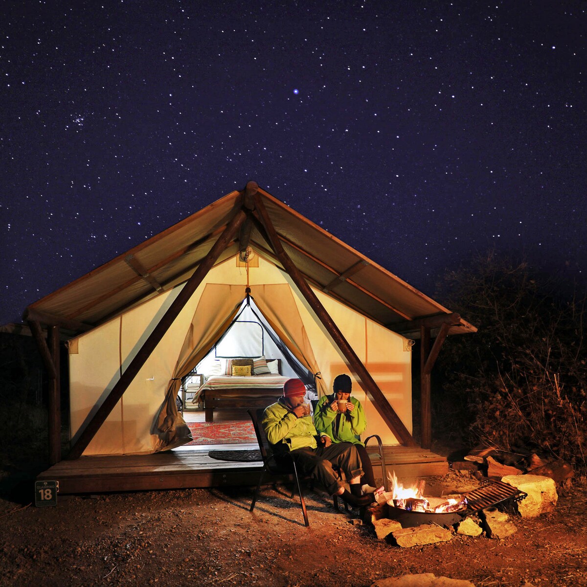 A glamping tent is framed by a clear night sky filled with stars. Two individuals sit in camping chairs outside, engaged by a fire pit. The tent entrance reveals a cozy interior with a bed visible inside, creating a warm ambiance.