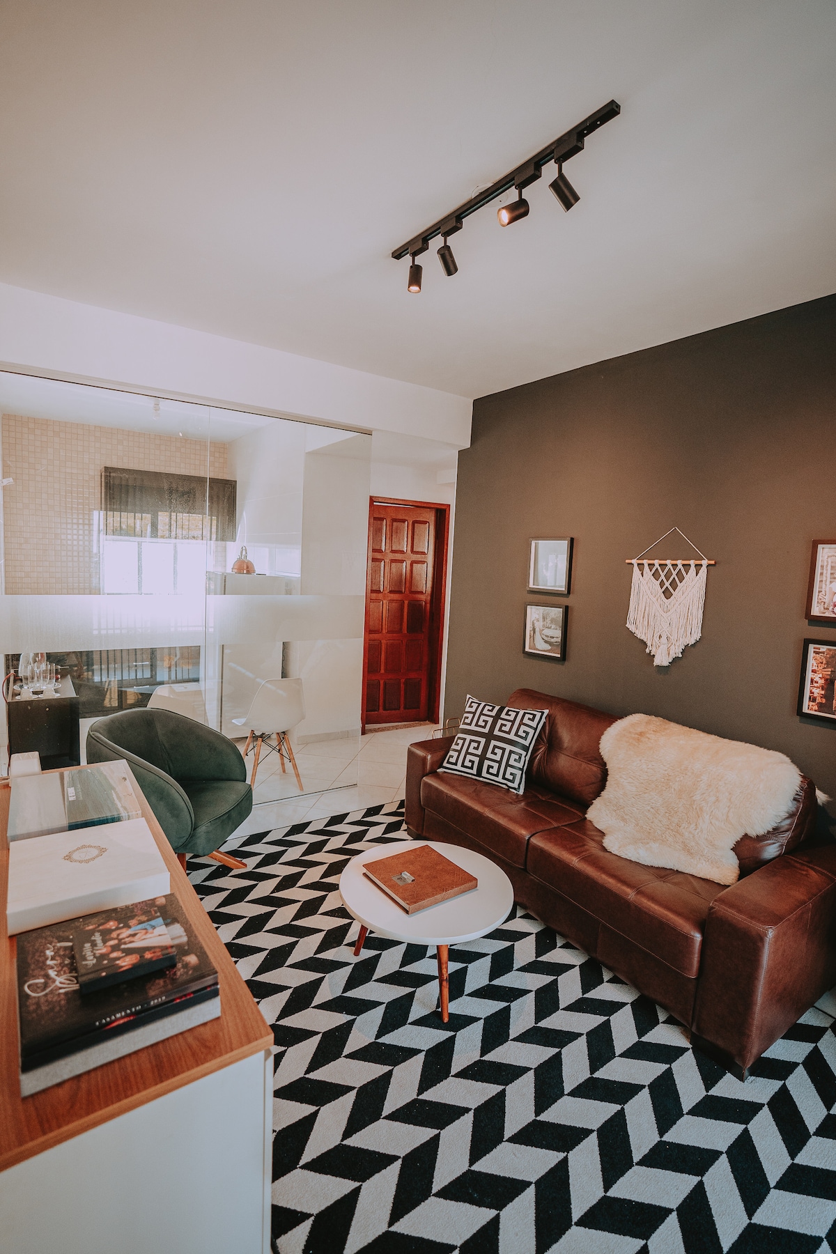 A living area is presented with a brown leather couch and a small round table. A patterned black and white rug covers the floor. Wall art and decorative items add character. Natural light is observed from the glass partition leading to the kitchen area.