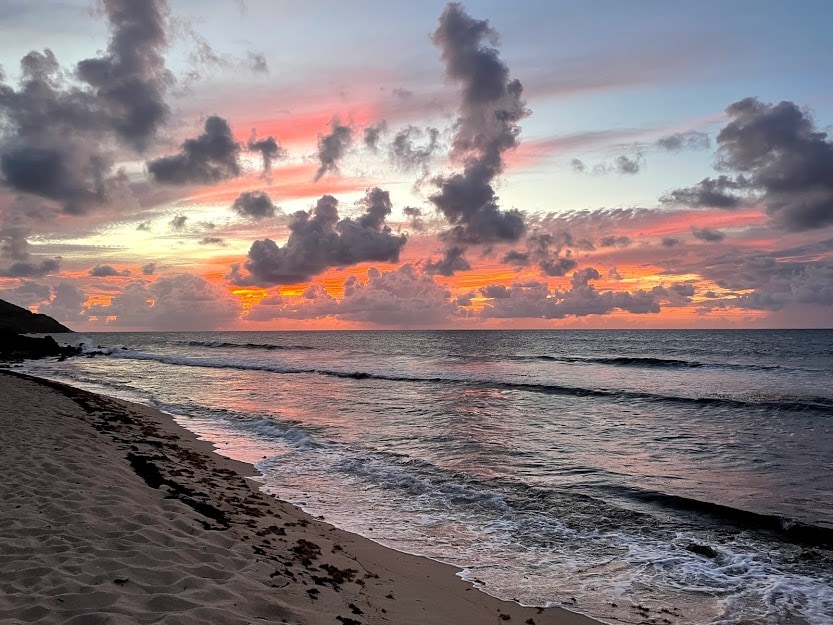 The shoreline is captured at sunset, showcasing gentle waves lapping against the sand. Vibrant hues of orange and pink fill the sky, creating a tranquil atmosphere, while clouds add depth to the scene. The shoreline reflects the colors, enhancing the coastal beauty.
